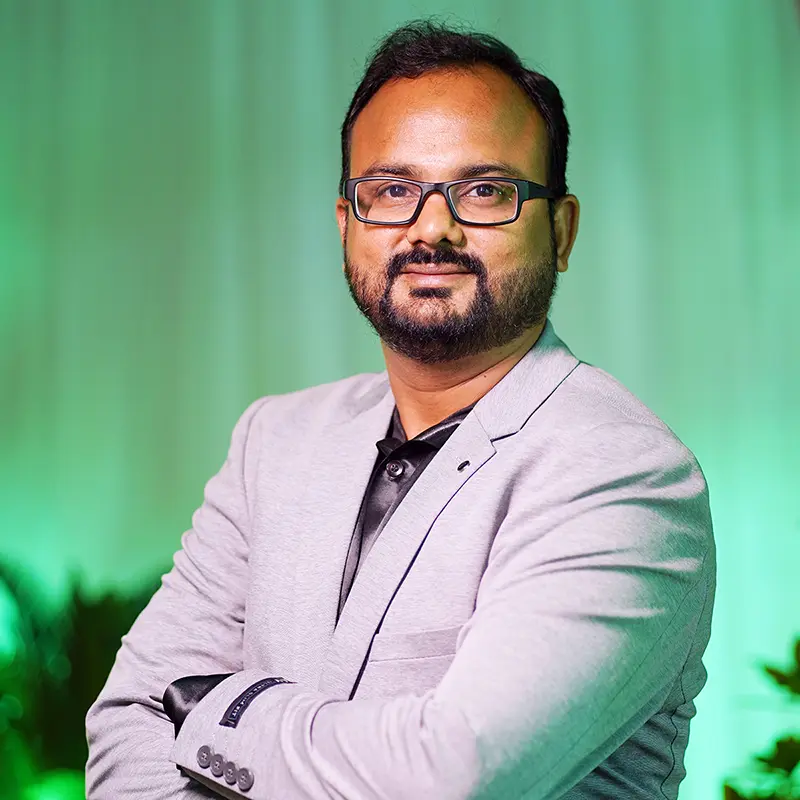Confident Indian man in grey blazer and glasses standing with arms crossed – professional headshot